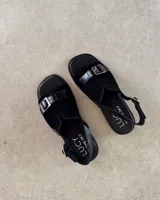 Sandalias negras con suela plana, tira ancha de gamuza con hebilla plateada y tira trasera ajustable.