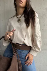 Blusa de color beige con diseño de corte holgado y mangas largas abullonadas. Presenta detalles bordados en tono sobre tono en las mangas y puños ajustados.