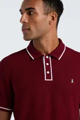 Chomba de piqué color bordó con detalles de ribetes blancos en cuello y mangas. Presenta cierre frontal de tres botones y logo de pingüino bordado en el pecho.