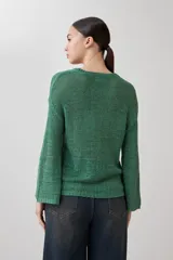 Sweater de punto fino en color verde, con cuello redondo y mangas largas. Presenta un detalle integrado de pañuelo estampado con motivos florales en la parte inferior, que se cruza sobre la cintura.