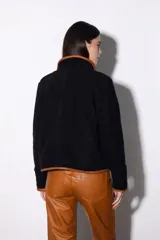 Chaqueta corta negra de paño con ribete de cuero color marrón en todo el contorno. Se ata con una tira de cuero en el centro y tiene dos bolsillos laterales.