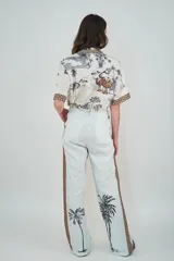 Pantalón de lino color celeste claro, de corte holgado y pierna recta. Presenta una franja lateral decorativa con estampado geométrico y detalles de palmeras estampadas en la parte inferior de las piernas.