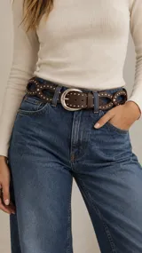 Cinto de cuero sintético marrón con diseño trenzado y tachas metálicas plateadas, hebilla ovalada y puntera clásica.