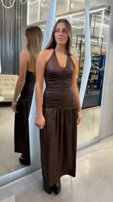 Vestido largo de fiesta color chocolate con acabado brillante. Presenta un diseño con escote halter, cuerpo ajustado con detalle de frunces centrales y falda fluida con corte a la cadera.