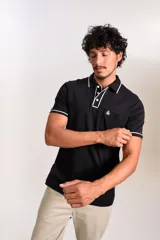 Polo de manga corta confeccionado en tejido interlock suave, color negro con detalles de vivos en color blanco en el cuello y las mangas. Presenta tapeta con botones y un bolsillo frontal con el logo bordado.