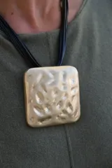 Collar de diseño con dije cuadrado de gran tamaño en tono dorado con textura irregular y martillada, suspendido de múltiples cordones negros finos.