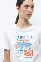Remera blanca de manga corta con cuello redondo y un estampado frontal que simula una barra de café o té con el texto "TEA D'CLOCK".