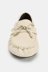 Mocasín plano de piel color beige, con diseño fruncido en la parte delantera, detalle de lazo frontal y cordón lateral entrelazado con ojalillos metálicos.