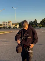Hoodie de color negro con diseño de estampa frontal que combina una tipografía manuscrita con el texto ITTAI STARS en letras mayúsculas. Presenta un corte holgado, capucha amplia y bolsillo tipo canguro.