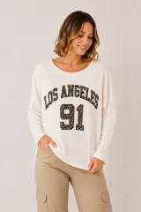 Buzo blanco de manga larga, con cuello redondo y diseño holgado. Presenta un estampado frontal con las palabras "LOS ANGELES" y el número "91" en animal print.