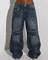 Pantalon de jean azul con diseño de corte holgado y estilo cargo. Presenta múltiples ojales metálicos decorativos dispuestos en franjas verticales y horizontales, con detalles de bordes deshilachados en las costuras.