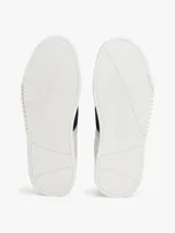 Zapatillas urbanas Tommy Hilfiger de corte bajo, confeccionadas en piel lisa color blanco. Presentan paneles laterales en azul marino y rojo, cordones oscuros y suela de goma gruesa con diseño dentado. Incluyen detalles de la marca en la lengüeta, lateral y talón.