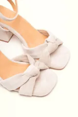 Sandalia de gamuza color beige con taco cuadrado, tira ajustable con hebilla en el tobillo y detalle de nudo en la tira delantera.
