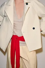 Trench corto color marfil de algodón, con cuello camisero, solapa cortaventos, mangas largas amplias con puños abotonados y cierre cruzado con un botón visible.