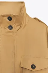 Chaqueta trench corta color beige, con cuello subido, manga larga con trabillas en hombros y mangas, solapas en pecho, bolsillos con solapa en delantero, cinturón lazada en mismo tejido, forro interior y cierre frontal con botones ocultos por solapa.