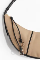 Bolso de hombro con forma de media luna, cuerpo principal en tela color beige y ribetes y correa en cuero sintético negro. Presenta cierre superior con tiradores largos de cordón negro anudado y argollas metálicas doradas.