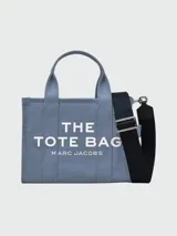 Bolso tote de lona en color azul con el texto 'THE TOTE BAG MARC JACOBS' estampado en blanco en el frente. Cuenta con asas de mano superiores y una correa de hombro ajustable y extraíble en color negro.