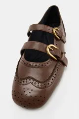 Zapato plano tipo bailarina de piel en color marrón, con detalles de perforaciones estilo brogue, doble tira delantera con hebillas metálicas doradas y puntera cuadrada.