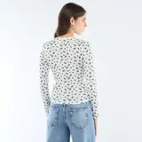 Camiseta de manga larga confeccionada en tejido de punto calado, con estampado de pequeñas flores azules sobre fondo blanco. Presenta un diseño ajustado al cuerpo, cuello redondo y una pequeña abertura frontal con botones.