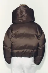Campera puffer corta de color marrón oscuro, con diseño acolchado, cuello alto y capucha integrada. Presenta cierre frontal con botones magnéticos y mangas largas con puños elásticos.