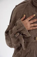 Chaqueta de corte estructurado con estampado cuadrille en tonos marrones y beige. Presenta cuello alto con tira ajustable, cierre frontal con botones, charreteras en los hombros y puños con detalle de hebilla.