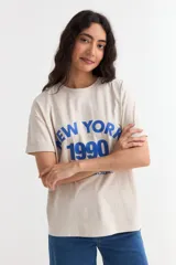 Remera de manga corta color beige, con estampa frontal en tono azul con la leyenda "New York 1990", de corte holgado y diseño casual.
