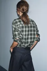 Camisa de corte oversize confeccionada en algodón, con estampado de cuadros en tonos verde, negro y blanco. Presenta cuello solapa, cierre frontal con botones, manga larga con puños y bajo asimétrico.