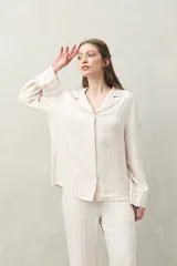 Camisa de pijama color blanco tiza con rayas verticales en relieve, de silueta clásica y caída fluida. Presenta cuello camisero, cierre frontal con botones y detalles de vivo rosado en cuello, puños y solapas.