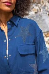 Camisa de denim color azul oscuro con efecto desgastado, corte oversize, manga larga y botones a presión. Presenta estrellas aplicadas con pedrería multicolor en todo el cuerpo de la prenda.