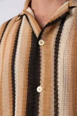 Camisa de manga corta con cuello solapa y cierre frontal de botones. Presenta un diseño de rayas verticales en tonos marrones, beige y negro, con una textura tejida y corte holgado.