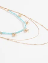 Collar triple de estilo delicado, compuesto por una cadena con cuentas celestes y dijes de flores plateadas, junto a dos cadenas simples de eslabones plateados.