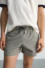 Bañador de hombre color verde claro con finas rayas blancas verticales (efecto seersucker). Tiene cintura elástica ajustable con cordón verde, bolsillos laterales y un bolsillo trasero de vivo.