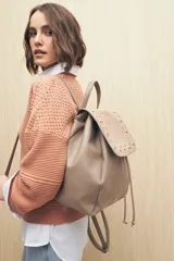 Mochila color visón de cuero sintético texturizado con solapa de gamuza sintética con tachas metálicas. Tiene asa superior, correas regulables y cierre con cordón ajustable.