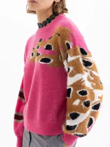 Sweater de punto color fucsia, con cuello redondo. Presenta un diseño de manchas abstractas en tonos marrón, negro y blanco en el frente y en la manga derecha. La manga izquierda tiene un diseño de rayas horizontales en tonos marrón y gris.