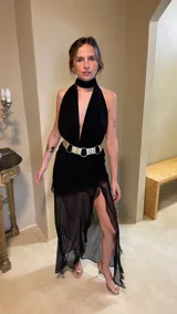 Vestido largo negro con escote profundo tipo halter y cuello alto. La falda presenta frunces y capas de microtul transparente. Se complementa con un cinturón ancho metálico de doble hebilla.