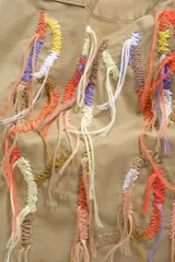 Bolso tipo shopper de color beige, con diseño amplio y doble asa de hombro. Presenta un detalle frontal de bordados coloridos con hilos colgantes que forman un diseño artístico.