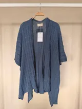 Ruana tejida de algodón con hilo lurex en punto jersey, color azul, con diseño amplio y detalle de etiqueta de cuerina en la parte posterior.