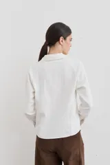 Remera de manga larga color blanco con cuello polo y cierre frontal parcial. Presenta costuras a la vista en contraste y un pequeño bordado en el pecho. El diseño incluye una abertura lateral en el ruedo.