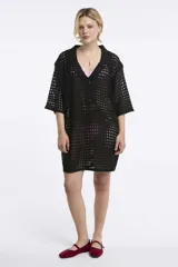 Vestido corto de tejido calado en color negro, con cuello polo, cierre frontal parcial con botones y mangas cortas. Presenta un diseño holgado y textura abierta.