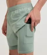Bermuda deportiva para hombre, color verde menta, con short de compresión integrado. La bermuda exterior es suelta y tiene un pequeño logo estampado en el muslo izquierdo.