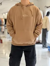 Canguro oversize beige con capucha y cordones ajustables, bolsillo tipo canguro y pequeño bordado en el pecho.