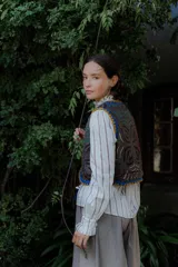 Chaleco de algodón color marrón oscuro con bordados en contraste de colores vibrantes como azul, amarillo, verde, rosa y blanco. Tiene bordes festoneados en azul y amarillo, y tiras para atar en el frente.