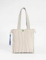 Cartera tipo shopper de color beige con diseño matelaseado de líneas onduladas y costuras en contraste azul. Cuenta con doble asa de hombro y un colgante decorativo de cordones multicolor en uno de sus laterales.