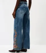 Pantalón wide leg de jean azul con bordado floral aplicado en los laterales. Posee cintura mediana con trabillas, cierre frontal con botón y cremallera, y bolsillos funcionales.