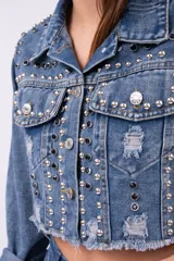 Campera de jean cropped con diseño deshilachado en el ruedo y detalles de roturas. Presenta una profusión de tachas metálicas plateadas y negras distribuidas por todo el frente, hombros y mangas, con cierre frontal de botones metálicos.
