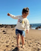 Sweater tejido a mano en lana merino color crudo con cuello redondo y mangas largas. Presenta la palabra "SURF" bordada en el frente con letras de colores.