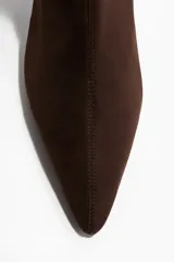 Botines tipo media de color marrón oscuro, con caña elástica ajustada, punta fina y taco bajo curvo forrado. Presentan costura central en la parte delantera.