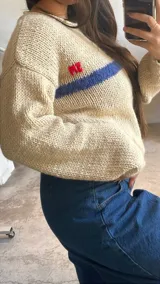 Sweater de punto en tonos beige con efecto degradado, cuello redondo y manga larga. Presenta una franja horizontal en color azul y el bordado de las letras 'SM' en color rojo en el pecho.