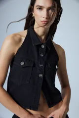 Chaleco de denim negro con escote halter, cuello camisero y cierre frontal con botones metálicos. Presenta dos bolsillos frontales con solapa y diseño sin mangas.
