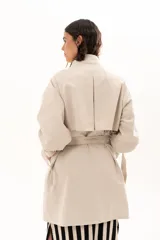 Trench corto color beige con cuello alto, cierre frontal de botones y cinturón ajustable en la cintura. Presenta mangas largas con detalles de tiras ajustables en los puños y bolsillos laterales.
